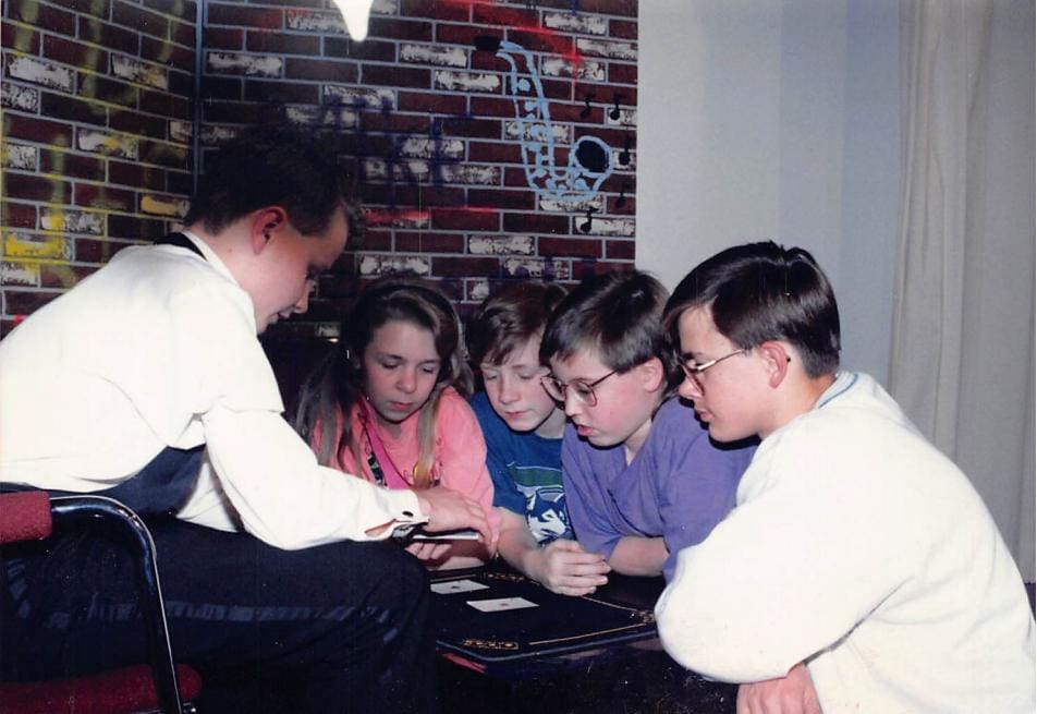 A teenage David Farr in shirt and vest performing card magic at a table for four young people leaning in to watch