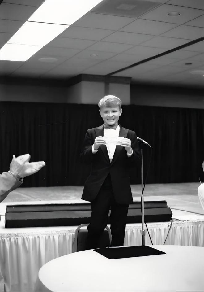A young David Farr in a tuxedo taking applause on stage at a microphone