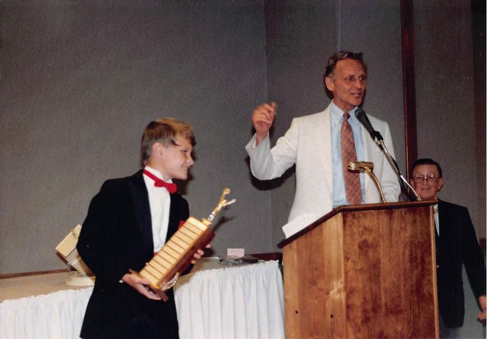 A young David Farr in a tuxedo accepting a tall gold trophy from a presenter at a podium