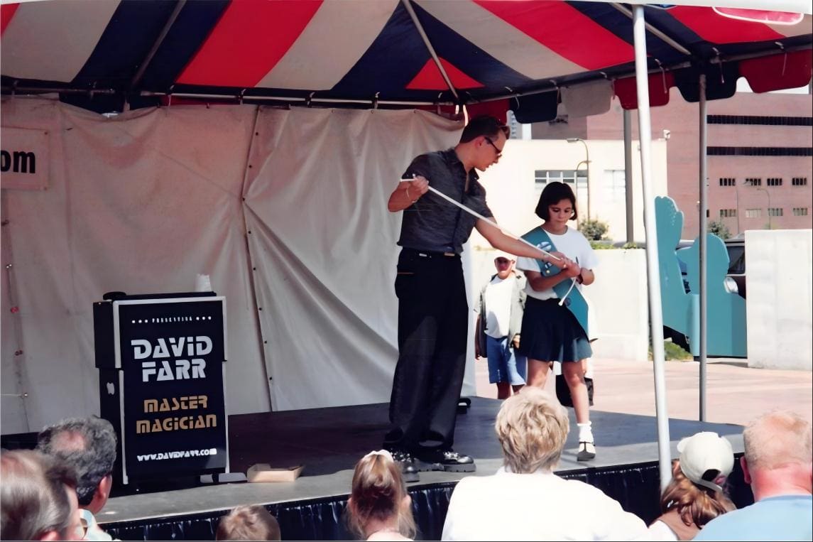 David Farr performing on an outdoor stage beneath a striped tent beside a sign reading David Farr Master Magician, with a young volunteer and a seated crowd